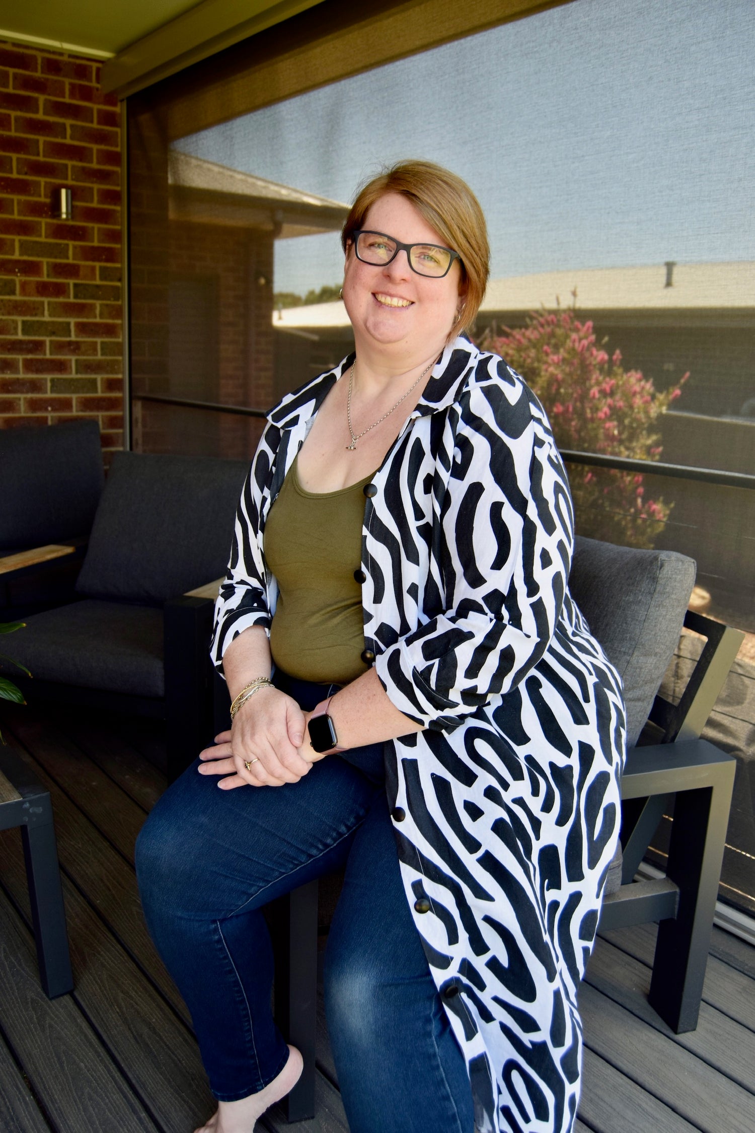 Woman sitting on a patio wearing a patterned coat, glasses, and a watch.