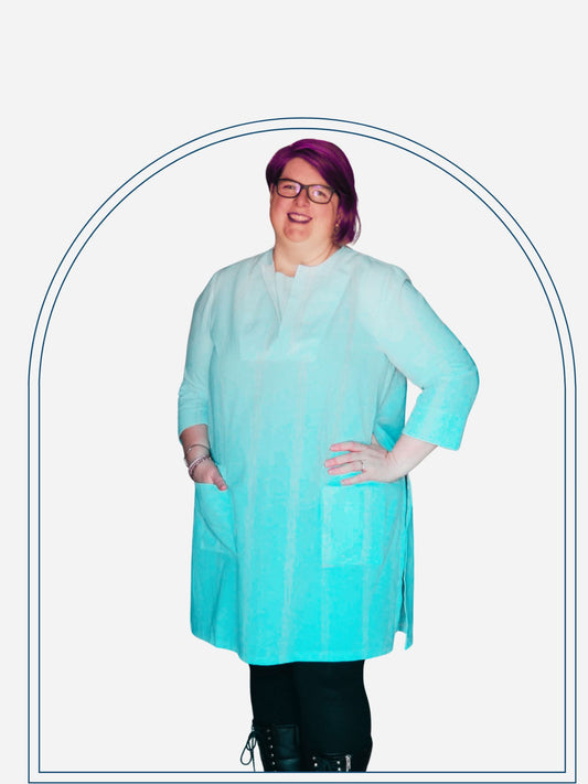 A woman standing in a doorway wearing a white tunic dress with 3/4 sleeves, paired with black boots and glasses.