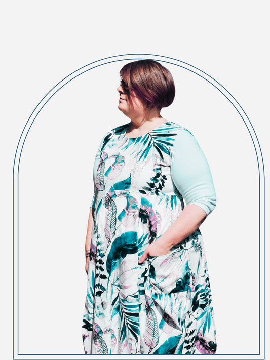 A woman wearing a floral print knee-length dress with sleeves, standing in front of a shrub.