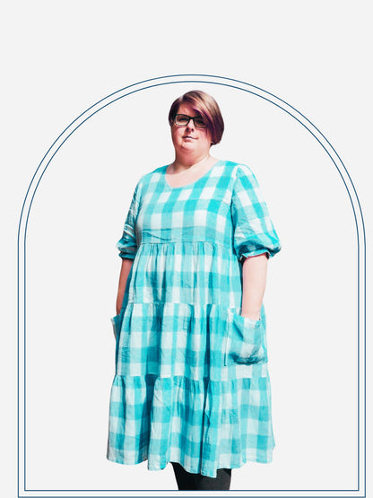 A person standing outdoors wearing a knee-length, short-sleeved, checkered tiered dress in purple and white, with pockets.