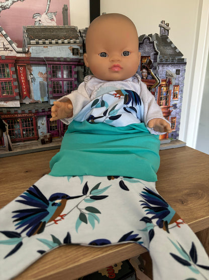 A doll wearing a blanket with a mermaid or shark tail design, positioned on a wooden table with a background of toy buildings and decorations.